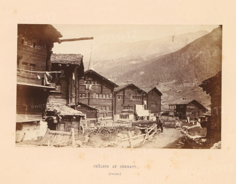 View of chalets in Zermatt, Châlets at Zermatt, photograph, page, inscription: '(valais)', photographer: Stephen Thompson, Zermatt, c. 1863 - in or before 1868, paper, albumen print, height 107 mm x width 176 mm, street (city(-scape) with figures, staffage), hut, cabin, lodge, A sepia-toned photograph showing a row of wooden chalets with steep shingled roofs and multiple small windows, built close together along a dirt path lined with simple wooden fences; a person is standing on the path near the foreground beside the fences; some cloth is hanging from a balcony or railing on one of the buildings; a tall spire rises behind the chalets and tree-covered mountains form the background under a pale sky.
