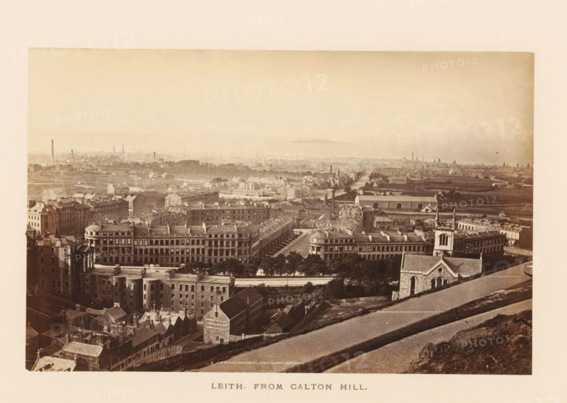 View of Leith, Leith, from Caltn Hill, photograph, page, photographer: Archibald Burns, Edinburgh, c. 1862 - c. 1867, paper, albumen print, height 154 mm x width 102 mm, Photo two pages after page 45., city-view in general, 'veduta', A wide sepia-toned panoramic view of a cityscape taken from an elevated vantage showing rows of multi-story stone buildings with pitched roofs and chimneys in the foreground, tree-lined streets and formal terraces of large rectangular buildings with repetitive windows and cornices, a church with a tower and arched windows near the right foreground, open grassy slopes and a sloping paved path in the lower right leading down toward the built area, densely packed residential blocks and narrow streets extending into a flatter middle ground with scattered industrial chimneys and low warehouses, expanses of lighter-toned open land and distant low hills on the horizon under a pale sky.
