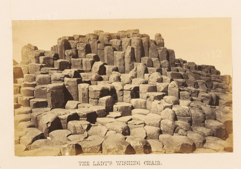View of rock formation the Lady's Wishing Chair, The Lady's Wishing Chair, photograph, page, photographer: John Hudson, Giant's Causeway, c. 1862 - in or before 1867, John Hudson, photographer, Causeway Coast and Glens, paper, albumen print, height 110 mm x width 178 mm, landscapes, rock-formations, A sepia-toned photograph showing a large area of interlocking, columnar rock formations with flat tops and vertical sides, arranged in terraces and clusters that rise toward the back of the image; the rocks display rough, weathered surfaces and edges with visible cracks and joints, and varying heights and shapes creating a stepped, geometric pattern across the scene, with a pale, empty sky above and a narrow pale border around the photograph.