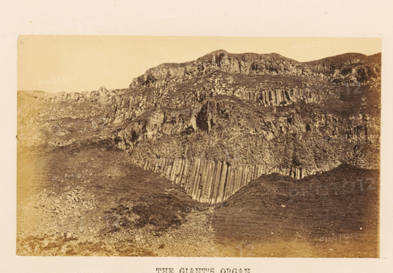 The Giant's Organ, View of rock formation The Giant's Organ, photograph, page, photographer: John Hudson, Giant's Causeway, c. 1862 - in or before 1867, John Hudson, photographer, Causeway Coast and Glens, paper, albumen print, height 109 mm x width 180 mm, landscapes, rock-formations, A sepia-toned photograph of a rocky hillside with layered, column-like rock formations and rough textured slopes, showing vertical striations and rugged cliff faces, with patches of sparse vegetation and scattered stones across the uneven ground, framed by a pale border.