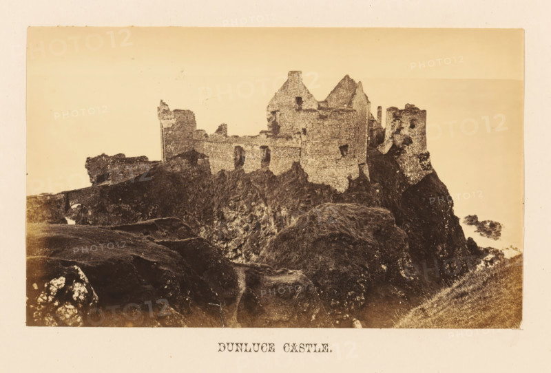 Dunluce Castle, View of Dunluce Castle, photograph, page, photographer: John Hudson, Giant's Causeway, c. 1862 - in or before 1867, John Hudson, photographer, Causeway Coast and Glens, paper, albumen print, height 108 mm x width 178 mm, castle, ruin of a dwelling, house, A sepia-toned photograph of a ruined stone fortress perched on a rocky promontory with crumbling walls and partially standing towers, showing rough stone textures, window openings, and jagged rooflines, surrounded by dark rocky cliffs and grassy slopes in the foreground and a pale, empty sky in the background.