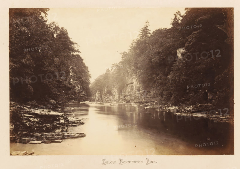 View of the Clyde below the Bonnington Linn, Below Bonnington Linn, photograph, page, South Lanarkshire, c. 1862 - in or before 1867, photographer, paper, albumen print, height 116 mm x width 179 mm, river, landscapes with waters, waterscapes, seascapes (in the temperate zone), A sepia-toned landscape photograph showing a calm river flowing between steep, rocky banks lined with dense trees and foliage; the river surface reflects light and the surrounding cliffs, with flat rock ledges and scattered boulders visible along the water’s edge, and tall trees rising above the cliffs on both sides under a pale sky.