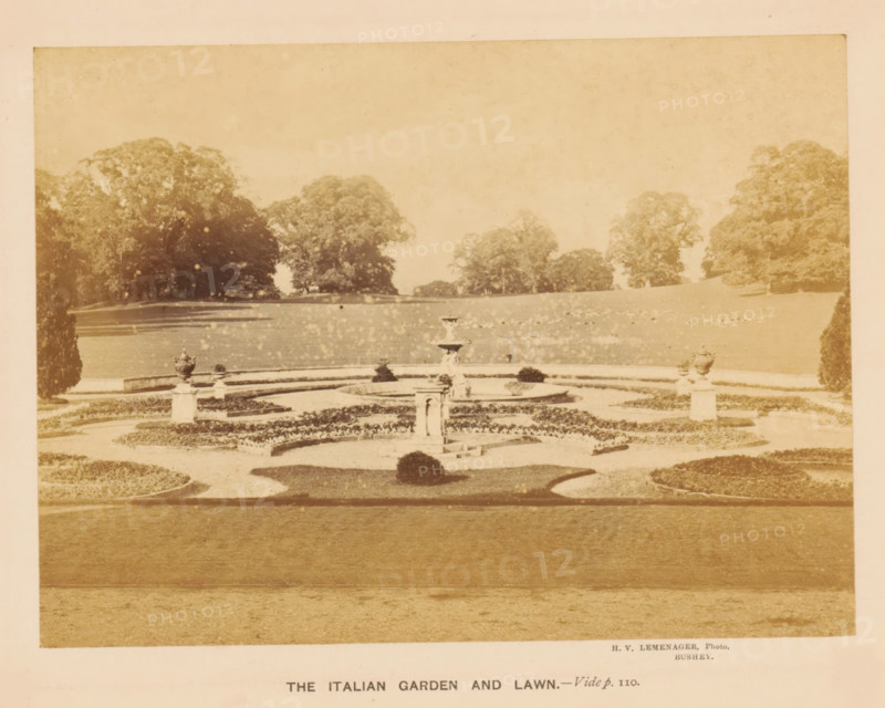 View of the Italian Garden in Moor Park in Rickmansworth, The Italian Garden and Lawn, photograph, page, inscription: 'Vide p. 110.', photographer: Henri Victor Leménager, Rickmansworth, c. 1866 - in or before 1871, Hertfordshire, paper, albumen print, height 103 mm x width 140 mm, park, garden ornaments, A faded sepia photograph of a formal garden with a central tiered fountain surrounded by concentric low hedges and gravel pathways, flanked by ornamental stone urns on pedestals, with symmetrical planted beds and clipped shrubs in the foreground and a wide open lawn leading to a distant line of large leafy trees under a pale sky.