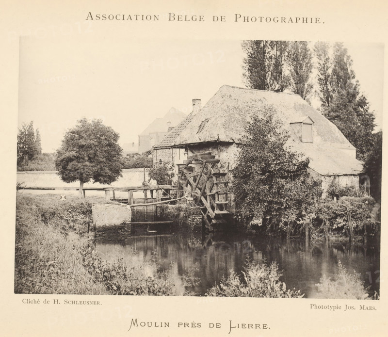 View of a watermill, Moulin près de Lierre, page, photomechanical print, inscription: 'Bulletin de l'Association belge de photographie', maker: Henri Schleusner, maker: Joseph Maes, c. 1885 - in or before 1890, Henri Schleusner, Joseph Maes, paper, collotype, photoprint, height 109 mm x width 155 mm, landscapes with waters, waterscapes, seascapes (in the temperate zone), watermill, A sepia-toned photograph shows a riverside scene with a thatched-roof building partially covered in climbing plants and foliage, a large wooden waterwheel attached to the building, a calm reflective body of water in the foreground, grassy banks and reeds along the water, a leafy tree near a simple wooden fence on the far bank, and tall trees rising behind the building.