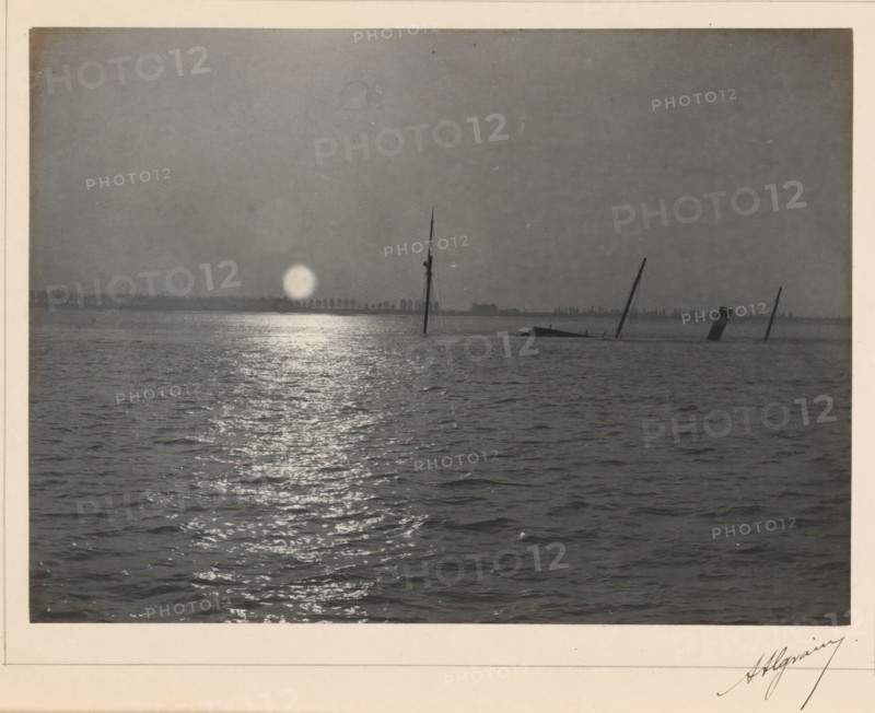 View of a sinking ship with a setting or rising sun, photograph, page, signature: 'Algrain', photographer: Alfred Algrain, c. 1885 - in or before 1890, paper, platinum process, height 158 mm x width 118 mm, landscapes with waters, waterscapes, seascapes (in the temperate zone), ships (in general), sun as celestial body, A grayscale photograph showing a wide expanse of water with rippling surface and a bright reflective path leading toward the horizon, where a low distant shoreline is visible. Near the horizon there are several partially submerged ship masts and a slanted smokestack emerging from the water, silhouetted against a pale circular light near the waterline. The sky above is plain and uniformly shaded, and the image is bordered by a light-colored matte.