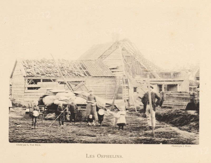 Children who were orphaned after an explosion of a munitions factory in Oosterweel near Antwerp in 1889, Les orphelins, page, photomechanical print, number: '13', photographer: L. van Neck, Oosterweel, cliché maker: anonymous, Belgium, printer (person): J. Maes (possibly), Antwerp, 1889, printer (person), paper, collotype, photoprint, height 165 mm x width 230 mm, Image on page 13., orphans, Explosion of a munitions factory in Oosterweel in 1889, A faded black and white scene shows a group of people standing and sitting in front of a cluster of wooden buildings with partially missing roof tiles and exposed rafters, scaffolding and ladders leaning against the structures, piles of sacks and covered carts in the yard, a man standing with a long pole, several children holding bowls or standing near baskets, rough grassy ground underfoot, and various wooden crates, fences, and farm equipment scattered around.