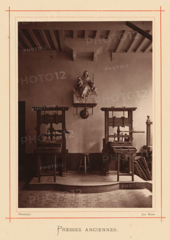 Presses anciennes, Printing presses in the house of Christoffel Plantijn, page, photomechanical print, maker: Joseph Maes, Antwerp, c. 1873 - in or before 1878, paper, Woodburytype, height 162 mm x width 117 mm, printing press, A framed sepia photograph showing an interior with a high beamed ceiling and a plain wall; mounted on the wall is a sculpted figure seated on a small pedestal with a round object hanging below it; below the sculpture are two tall wooden presses positioned symmetrically with a small wooden stool centered between them; the floor in front of the raised platform is made of rectangular tiles and the platform has a curved front edge; stacks of flat boards or frames lean against the wall near one press; the photograph is bordered by a cream mount with thin red lines near the edges.