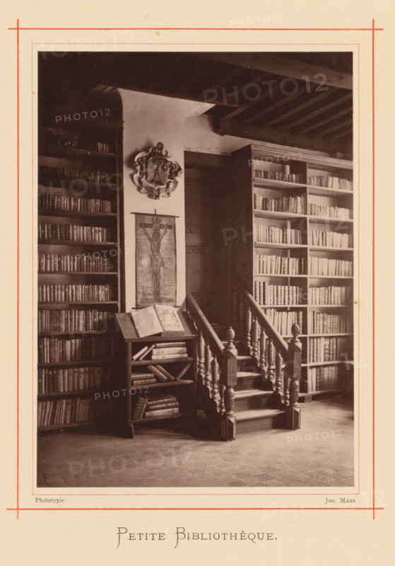 Interior of the library in the house of Christoffel Plantijn, Petite bibliothèque, page, photomechanical print, maker: Joseph Maes, Antwerp, c. 1873 - in or before 1878, paper, Woodburytype, height 164 mm x width 118 mm, study, 'studiolo', library, A sepia photograph shows an interior with tall bookshelves filled with books on either side of a wooden doorway, a carved wooden staircase with turned balusters and newel posts leading up beside the doorway, a small open bookrest holding an open book and additional books on its shelves, a framed image of a crucifix hanging on the wall between the shelves, a decorative crest mounted above the image, exposed wooden ceiling beams overhead, and a tiled floor in the foreground.