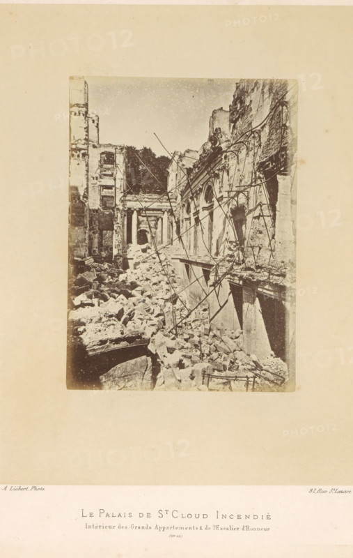 View of the remains of the grand rooms and grand staircase in the Château de Saint-Cloud after the fire caused by the Paris Commune, Le Palais de St. Cloud Incendié. Intérieur des Grands Appartements & de l'Escalier d'Honneur, photograph, page, number: '(No. 61)', address: '31, Rue St. Lazare', photographer: Alphonse-Justin Liébert, Saint-Cloud, 1871 - 1872, paper, albumen print, height 136 mm x width 99 mm, stairwell, ruin of a building, architecture, castle, Paris Commune (1871), Château de Saint-Cloud, A sepia-toned photograph shows a ruined interior of a large stone building with collapsed floors and piles of rubble filling the ground level, exposed masonry and broken arches along one side, tall fractured walls with empty window openings and jagged edges, scattered beams and twisted metal rods leaning and entangled across the scene, distant remains of columns and an archway partially visible toward the back, irregular stones and debris strewn across the foreground, and a pale blank...