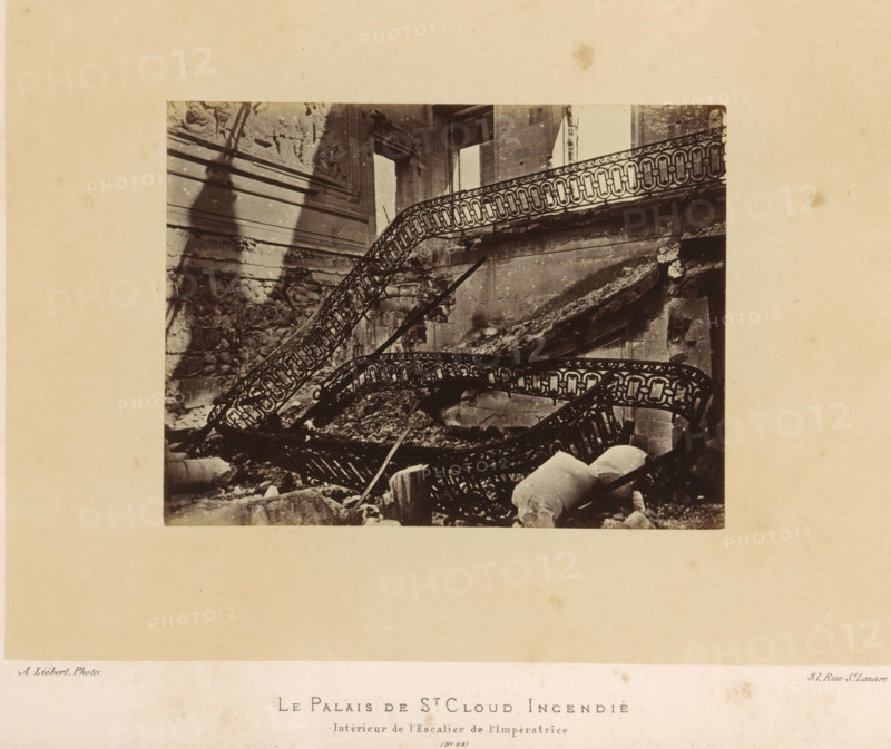 The Palace of St. Cloud Burned. Interior of the Empress's Staircase, View of the Empress's staircase in the Château de Saint-Cloud after the fire caused by the Paris Commune, photograph, page, address: '31, Rue St. Lazare', number: '(No. 62)', photographer: Alphonse-Justin Liébert, Saint-Cloud, 1871 - 1872, paper, albumen print, height 99 mm x width 131 mm, stairwell, ruin of a building, architecture, castle, Paris Commune (1871), Château de Saint-Cloud, A sepia photograph shows a ruined interior with twisted ornate metal stair railings collapsing amid rubble and broken stone fragments, a partially intact carved wall with decorative reliefs and moldings on the left, a large rectangular window opening with shutters on the right, crumbling masonry and fallen architectural elements scattered on the floor, and a pale surrounding border framing the image.
