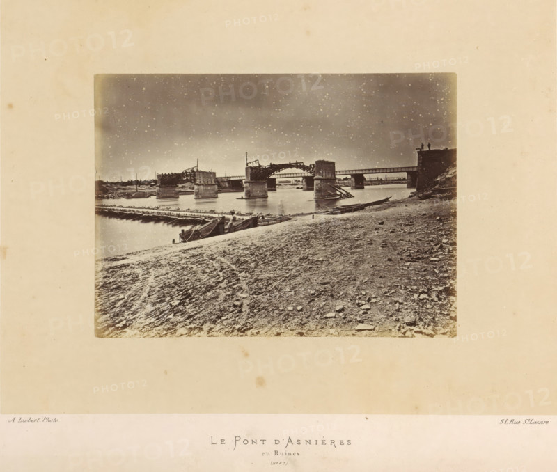 View of the remains of the bridge of Asnières-sur-Seine after the destruction by the Paris Commune, Le Pont d'Asnières and Ruines, photograph, page, number: '(No. 67)', address: '31, Rue St. Lazare', photographer: Alphonse-Justin Liébert, Asnières-sur-Seine, 1871 - 1872, Alphonse-Justin Liébert, photographer, paper, albumen print, height 99 mm x width 135 mm, ruin of a building, architecture, bridge, Paris Commune (1871), A sepia-toned photograph showing a riverbank foreground with a textured, rocky shore and scattered stones, a few wooden boats drawn up at the water's edge, and a timber walkway or floating bridge extending across the water toward a partially ruined bridge structure with several stone piers and damaged wooden arches; the sky appears mottled and grainy above the bridge, and two small human silhouettes stand on a raised platform or abutment at the far right of the bridge.