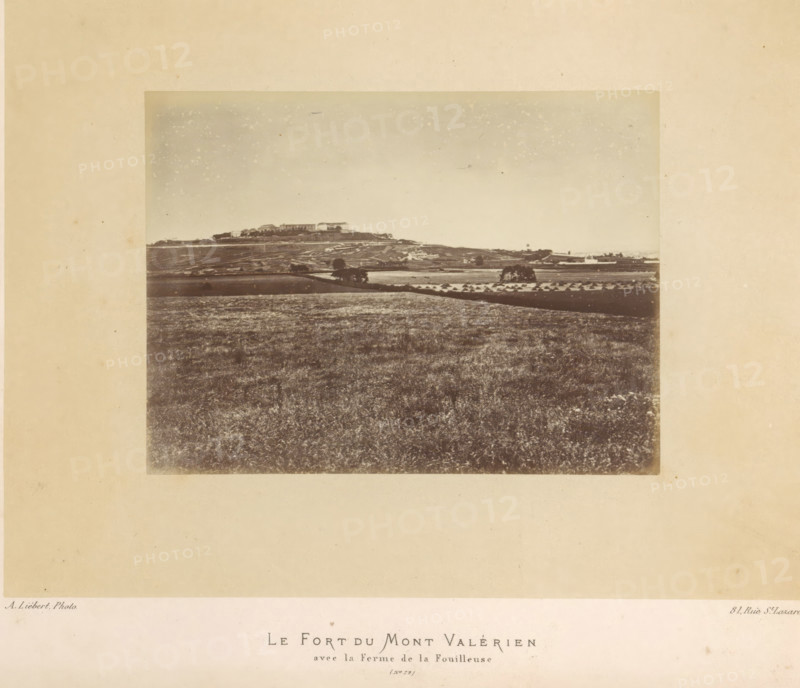 View of the fortress of Mont-Valérien in Suresnes, Le Fort du Mont Valérien avec la Ferme de la Fouilleuse, photograph, page, number: '(No. 72)', address: '31, Rue St. Lazare', photographer: Alphonse-Justin Liébert, Suresnes, 1871 - 1872, paper, albumen print, height 99 mm x width 132 mm, fortress, Paris Commune (1871), Mont Valérien, A sepia-toned photograph mounted on a wide beige matte shows a distant hilltop with a cluster of rectangular buildings and lower structures along its crest, a patchwork of fields and terraces spread across the slope and foreground, a few isolated trees and small groves dotting the midground, rows of small conical haystacks visible in one field, and a broad expanse of textured grassy land filling the lower portion of the image under a pale, lightly speckled sky.
