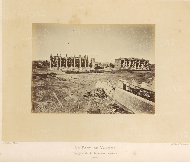 View of Fort de Vanves in Vanves after the destruction by the Paris Commune, Le Fort de Vanves. Vue Générale du Panorama intérieur, photograph, page, number: '(No. 78)', address: '31, Rue St. Lazare', photographer: Alphonse-Justin Liébert, Vanves, 1871 - 1872, paper, albumen print, albumendruk, height 99 mm x width 135 mm, ruin of a building, architecture, fortress, Paris Commune (1871), Fort de Vanves, A sepia-toned photograph of a wide open area with ruined buildings along the horizon, showing two large multi-story structures with exposed interiors and damaged facades, piles of rubble and debris scattered in front of them, a flat foreground of dirt and uneven ground with scattered fragments and low walls, and a partially collapsed structure with visible beams and rubble in the lower right corner under a sky lightly speckled with spots.