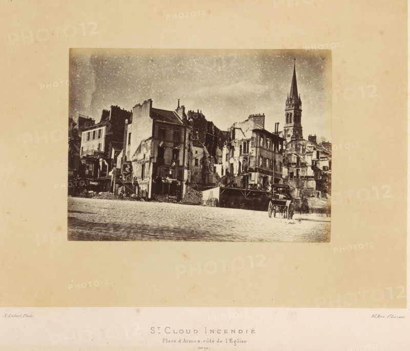 St. Cloud Incendié. Place d'Armes, côté de l'Eglise, View of the Place d'Armes in Saint-Cloud after the fire by the Paris Commune, photograph, page, number: '(No. 56)', address: '31, Rue St. Lazare', photographer: Alphonse-Justin Liébert, Saint-Cloud, 1871 - 1872, Alphonse-Justin Liébert, photographer, paper, albumen print, height 98 mm x width 133 mm, square, circus, ruin of a building, architecture, Paris Commune (1871), A sepia-toned photograph shows a row of damaged multi-story buildings with exposed interiors, crumbling facades, and rubble at their bases, with a tall church steeple rising behind them; a cobbled open area is in the foreground where a two-wheeled cart is parked, and the sky above the buildings is grainy with scattered light spots.