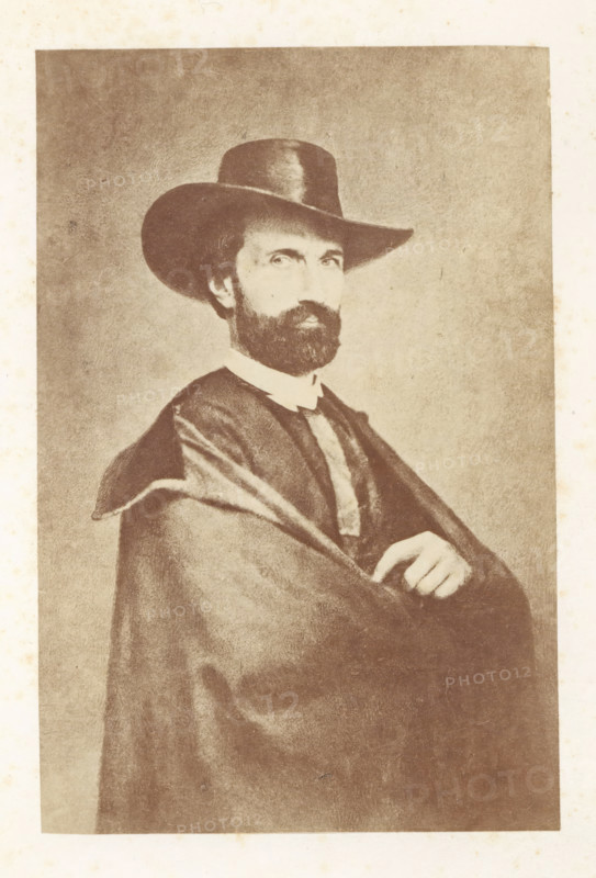 Portrait of Antoine Wiertz, photograph, page, check stamp: 'SOCIÉTÉ ROYALE BELGE DE PHOTOGRAPHIE, 69, RUE KEYENVELD, BRUXELLES', photographer: Edmond Fierlants, c. 1863 - in or before 1865, paper, albumen print, height 161 mm x width 106 mm, Person, Antoine Joseph Wiertz, historical persons, portrait, self-portrait of painter, A sepia-toned portrait of a bearded man wearing a wide-brimmed hat and a dark cloak draped over his shoulders, showing his crossed arms and one hand resting near his chest; his face is turned slightly toward the viewer, with visible eyes and mustache, and the background is a plain textured surface.