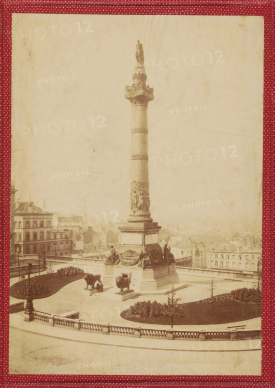 View of the Congress Column in Brussels, photograph, page, Brussels, c. 1854 - in or before 1859, photographer, paper, albumen print, height 155 mm x width 109 mm, Photo on front cover, column, pillar, architecture, Congress column, A tall stone column topped by a standing statue rises from a square base adorned with sculpted figures, with two large lion statues positioned on pedestals near the base; the column and base are set within a paved terrace bordered by a balustrade and low curved walls, with manicured lawns and planted beds visible in the surrounding area; several multi-story buildings with rows of windows and pitched roofs are visible in the background; a few people are seen near the monument and on the terrace; the image has a faded, sepia tone and is framed by a patterned red border.