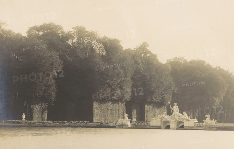 Le Lac et le Groupe de Neptune, View of a lake containing sculptures of Neptune, photograph, page, photographer: Coppin-Goisse, c. 1904 - in or before 1909, paper, gelatin silver print, developing gelatin silver print, height 83 mm x width 126 mm, sculpture, lake, Beloeil, A wide rectangular photograph bordered by a lighter mat shows a body of water in the foreground with a sculptural fountain group on a stone platform near the shore, featuring multiple classical figures and reclining horse forms, and a standing central figure holding a staff. Behind the fountain a row of closely planted, tall, trimmed trees forms a dense leafy screen with sculpted archways or openings at their bases. The sky above the trees is pale and largely empty, and the photograph is set against a brown textured backing with a thin painted rectangular border near its edges.