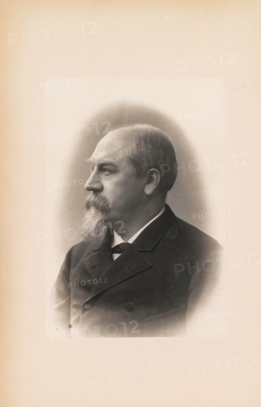 Portrait of Édouard Remouchamps, photograph, page, c. 1883 - in or before 1888, photographer, paper, platinum process, height 141 mm x width 100 mm, Photo opposite title page., Person, Édouard Remouchamps, historical persons, A sepia-toned portrait of an older man shown in profile facing left, with a receding hairline and short hair, a thick mustache and a long pointed beard, wearing a dark formal jacket with a visible collar and a tie, set against a softly shaded oval vignette on a pale background.