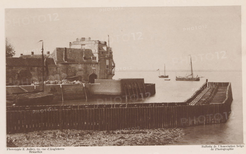 View of a dock, page, photomechanical print, inscription: 'Bulletin de l'Association belge de Photographie', inscription: 'Cliché au Gélatino-bromure', photographer: Hector Colard, cliché maker: Emile Aubry, 1878 - in or before 1883, Hector Colard, Emile Aubry, photographer, cliché maker, paper, collotype, photoprint, height 106 mm x width 179 mm, Print two pages before page 66., dry-dock, floating-dock, A sepia-toned harbor scene shows a weathered stone building with a tiled roof and visible chimneys situated on a quay, with an arched opening at the waterline and an external staircase on one side; wooden pilings and a curved timber breakwater extend into calm water in front of the building, and small sailing vessels with masts and rigging are moored a short distance offshore, one flying a small flag; the foreground shows a pebble-strewn shoreline and rows of vertical wooden posts forming a barrier, while the sky above is featureless and light in tone.