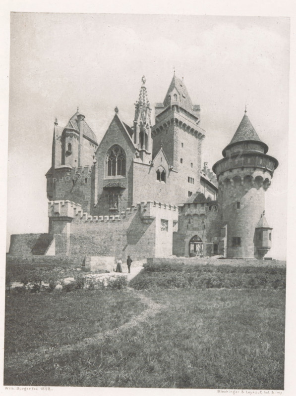 View of Burg Kreuzenstein, Burg Kreuzenstein, page, photomechanical print, photographer: Wilhelm Burger, cliché maker: Blechinger & Leykauf, c. 1894 - in or before 1899, printer (person), paper, photoaquatint, height 121 mm x width 91 mm, castle, A large stone fortress with multiple towers and turrets occupies the upper portion of the image, featuring crenellated walls, arched windows, and a prominent circular tower with a conical roof on the right; a central section shows a large pointed window and ornate stonework including a decorative pinnacle; a fortified gate with a portcullis and an adjacent smaller turret are visible near the circular tower; in front of the fortress a low wall and battlements run along the edge of a grassy area where two small human figures stand on a dirt path leading toward the entrance; the foreground consists of an open grassy field with a faint winding trail.