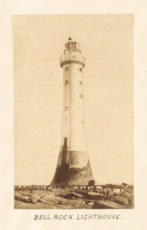 Bell Rock Lighthouse, View of the Bell Rock lighthouse, photograph, page, Scotland, c. 1867 - c. 1872, photographer, paper, albumen print, height 173 mm x width 119 mm, sea, lighthouse, lightship, A tall cylindrical lighthouse rising from a broadened base with visible texture and weathering, a narrow vertical seam running up its shaft, small rectangular windows set at intervals, a balcony with a railing near the top, and a glassed lantern room with a small finial above; rocky ground and a low wooden fence appear at the base, with a small structure near the lighthouse and scattered stones in the foreground.