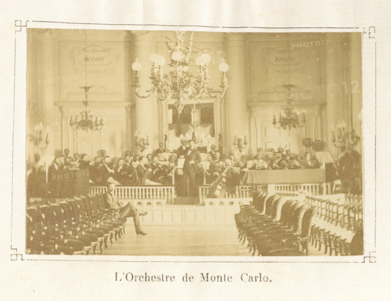 Les jeudis de Monte Carlo : impressions musicales, Ch.M. Domergue, book, dedication: 'à mon... le C... Barreme, souvenir ... de l'author, Domergue', date: 'JAN, 2003', bookplate: 'BIBLIOTHÈQUE du CHATEAU de BEAUREGARD', writer (clerical worker): Charles Mathieu Domergue, 1875, Charles Mathieu Domergue, writer (clerical worker), paper, printing, albumen print, height 188 mm x width 108 mm x thickness 20 mm, A sepia-toned photograph of an ornate interior showing rows of upholstered chairs arranged along a central aisle leading to a raised platform where a group of musicians are seated and standing with instruments, a conductor visible near the front and a large string instrument resting at the edge of the platform; the room features tall columns, decorative wall panels, multiple chandeliers hanging from the ceiling, wall-mounted candelabra, and framed artwork or mirror behind the musicians, with a single seated man visible in the aisle near the foreground.