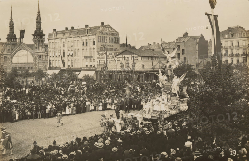 Wagon, glorification of Antoon van Dijck, photograph, page, Antwerp, c. 1899, photographer, paper, printing, height 98 mm x width 152 mm, civic architecture, edifices, dwellings, animal traction, animal-drawn vehicle (carriage etc.), festivities (decorated float, wagon, festive activities), A crowded street scene with a large procession float pulled by teams of horses and adorned with statues and figures, surrounded by a dense crowd of people wearing period clothing and hats, buildings with ornate facades and tall spires in the background, trees partially framing the scene, and lamp posts and banners visible above the assembled spectators.