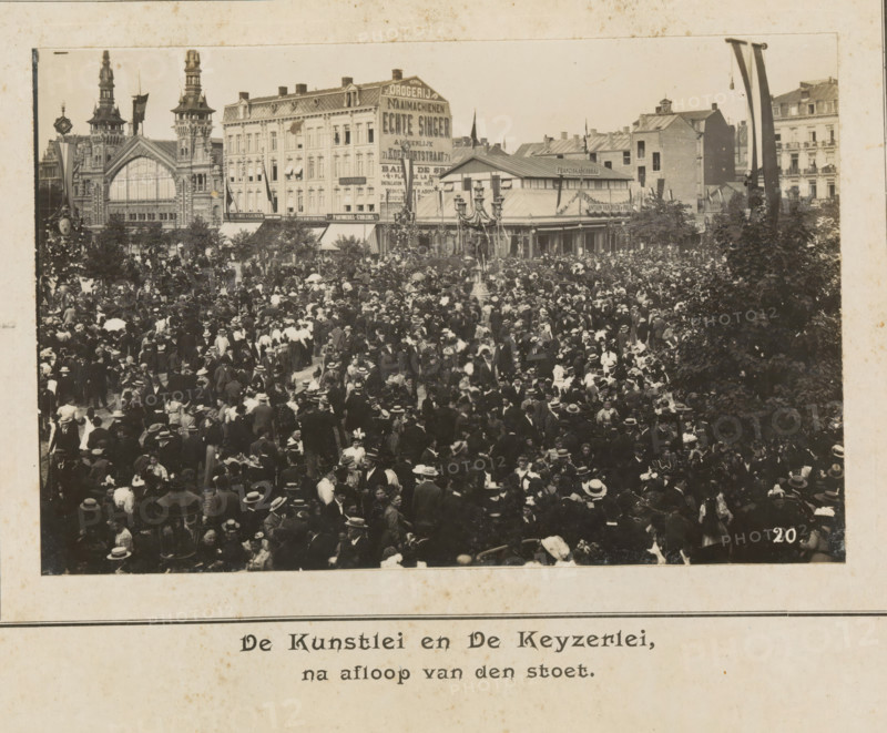 The Kunstlei and the Keyzerlei, after the procession, photograph, page, Antwerp, c. 1899, photographer, paper, printing, height 98 mm x width 152 mm, civic architecture, edifices, dwellings, festivities (decorated float, wagon, festive activities), A sepia-toned photograph showing a densely packed outdoor crowd with people wearing period clothing and hats, occupying a square in front of a row of ornate multi-story buildings with decorative facades and tall towers, a large arched window in one building, and a covered pavilion with a pitched roof. A tall ornate street lamp stands amid the crowd, trees or large bushes frame parts of the scene, and flags or long banners hang from poles near the buildings. The crowd includes men and women in dark and light garments, many holding parasols or wearing wide-brimmed hats, with groups clustered closely together across the foreground and middle distance.