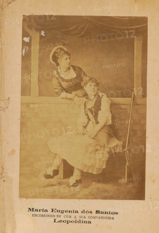 Portrait of Maria Eugenia dos Santos and an unknown woman, Maria Eugenia dos Santos, photograph, page, inscription: 'ENCOBRINDO-SE COM A SUA COMPANHEIRA, Leopoldina', c. 1881 - in or before 1886, photographer, paper, albumen print, height 140 mm x width 96 mm, Photo after cover page., Person, historical persons - BB - woman, A faded sepia photograph mounted on aged paper shows two women in period costumes posed by a low brick wall and a curtained backdrop. One woman stands behind the wall, leaning on it with one arm and looking to the side; she wears a dark bodice with decorative trim, a light blouse with elbow-length sleeves, a necklace, and a headpiece with a circular halo-like element. The other woman sits on the wall edge, facing slightly forward with hands resting in her lap; she wears a layered skirt with ruffled hem, a vest over a blouse, stockings, and shoes, and a necklace. A long slender object rests upright against the wall beside the seated woman. The photograph has worn ed...