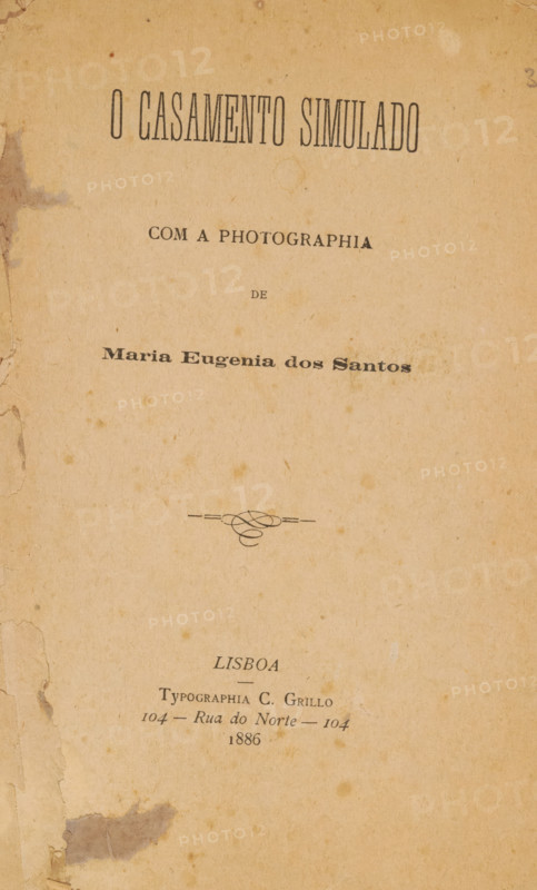 O casamento simulado com a photographia de Maria Eugenia dos Santos, book, typographer: C. Grillo, Lisbon, 1886, paper, printing, albumen print, height 228 mm x width 141 mm x thickness 6 mm, A vertically oriented aged paper page with a warm tan tone and scattered darker spots and stains, edges showing wear and small tears with a larger darker stain near the lower left margin, a decorative flourish centered above the lower third, and multiple areas of uneven discoloration across the surface.