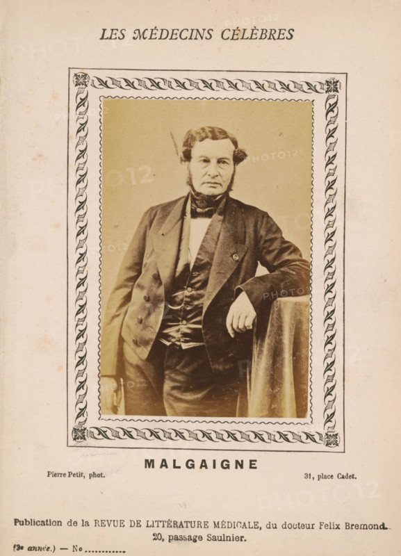Malgaigne, Portrait of Joseph-François Malgaigne, photograph, page, inscription: '(Publication de la Revue de literary médicale, du docteur Félix Bremond, 20, Passage Saulnier, Paris., 3e année - No.', photographer: Pierre Petit, c. 1871 - c. 1879, paper, albumen print, height 89 mm x width 56 mm, Person, Joseph-François Malgaigne, historical persons, adult man, A sepia-toned portrait of a man with muttonchop sideburns and a formal dark suit, standing with one arm resting on a draped surface, framed by a decorative border with a repeating leaf motif; the image shows the man's jacket, waistcoat, bow tie, and a visible ring on his hand, set against a plain backdrop and mounted on aged paper with faint spots and discoloration.