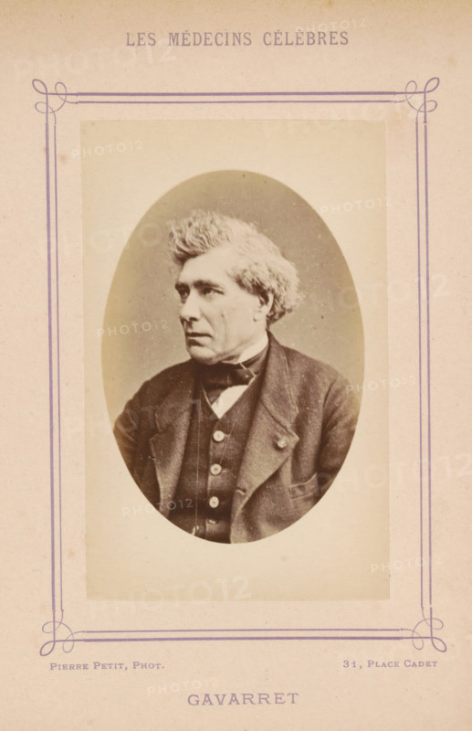 Portrait of Louis Denis Jules Gavarret, Gavarret, photograph, page, inscription: '(Publication de la Revue de literature médicale, du, DF Bremond, 20, Passage Saulnier, Paris., No.', photographer: Pierre Petit, c. 1871 - c. 1879, paper, albumen print, height 90 mm x width 56 mm, Person, Louis Denis Jules Gavarret, historical persons, adult man, An oval black-and-white portrait of an older man mounted on a faded beige card with a decorative purple border, showing the man from the chest up in three-quarter profile facing left, with wavy light-colored hair, a serious expression, a dark jacket over a buttoned vest and a bow tie, and faint blemishes and age marks visible on the card and photograph.