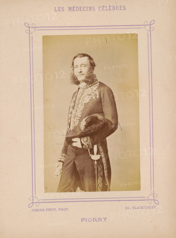 Portrait of Piorry, Piorry, photograph, page, inscription: 'Publication de la Revue de literature médicale, du, ... ) - No.', photographer: Pierre Petit, c. 1871 - c. 1879, paper, albumen print, albumendruk, height 91 mm x width 56 mm, historical persons, adult man, A faded sepia portrait of a man standing three quarters turned, wearing a dark ornate coat with embroidered floral and leaf patterns and decorative cuffs, holding a fur-trimmed hat under one arm and a pair of gloves in one hand, with prominent sideburns and short hair, framed by a simple rectangular border printed on aged paper.