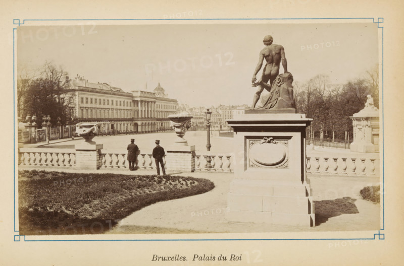 View of the Royal Palace in Brussels, Bruxelles. Palais du Roi, photograph, page, Brussels, c. 1875 - c. 1880, photographer, paper, albumen print, height 101 mm x width 175 mm, monument, statue, palace, Koninklijk Paleis (Brussel), A sepia-toned photograph showing a formal exterior scene with a large stone balustrade and decorative urns, a paved walkway curving past a planted lawn, and two men in dark coats and hats standing near the railing facing away from the camera; a tall stone pedestal topped with a muscular nude male statue seen from behind stands prominently to the right, draped fabric carved beside the figure, and an ornate rectangular panel on the pedestal; behind the balustrade a wide open paved forecourt stretches toward a long classical building with rows of windows and columned portico, trees and garden elements are visible at the edges of the scene, and a freestanding lamppost stands near the center distance.