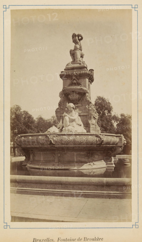 Bruxelles. Fontaine de Brouckère, View of the Charles de Brouckère fountain in Brussels, photograph, page, Brussels, c. 1875 - c. 1880, photographer, paper, albumen print, albumendruk, height 171 mm x width 97 mm, ornamental fountain, A large ornate stone fountain sits in a shallow pool with a circular basin decorated with carved shell and floral motifs and sculpted animal heads at intervals, a seated female figure in draped clothing rests on the raised central pedestal surrounded by other sculpted figures and decorative reliefs, above them a smaller group of figures is topped by a standing figure holding a wreath, trees rise in the background and paved stone surrounds the fountain.