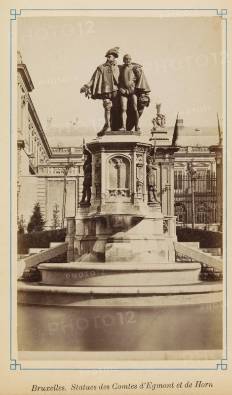 Brussels. Statues of the Counts of Egmont and Horn, Statue of Egmont and Horn on the Petit Sablon in Brussels, photograph, page, Brussels, c. 1875 - c. 1880, photographer, paper, albumen print, height 171 mm x width 102 mm, Person, Philip of Montmorency (Count of Horne), Lamoraal Count of Egmond, monument, statue, ornamental fountain, A sepia-toned photograph of a large ornate stone fountain topped by two standing male statues dressed in historical attire, one with a feathered hat and the other holding a hat at his side, both draped in capes and positioned close together on a high pedestal carved with decorative panels and reliefs; the fountain basin in front shows smooth rounded tiers and water flowing into the lower pool, while the pedestal sides feature additional sculpted figures in niches and ornamental motifs; behind the monument a classical building facade with columns, windows, balustrades, and sculptural roof details is visible, framed on the left and right by trimmed shrubs a...