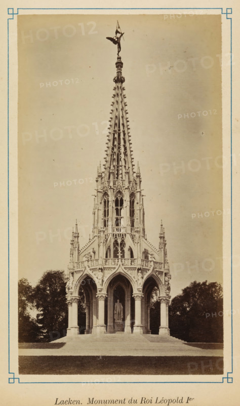 View of the Monument to the Dynasty, Laeken. monument du Roi Léopold 1er, photograph, page, Laeken, c. 1875 - c. 1880, photographer, Brussels, paper, albumen print, albumendruk, height 172 mm x width 98 mm, monument, statue, A tall ornate stone monument with a pointed spire and intricate Gothic detailing stands on a stepped base surrounded by trees, the lower section featuring arched openings supported by columns and a statue visible inside one of the arches, the spire topped with a sculpted figure and decorative finials, all set against a plain sky and framed by a light border.
