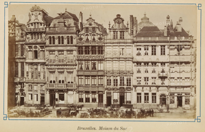 Brussels. Maison du Sac, View of the house Den Sack in Brussels, photograph, page, Brussels, c. 1875 - c. 1880, photographer, paper, albumen print, height 103 mm x width 172 mm, street (city(-scape) with figures, staffage), façade (of house or building), A row of ornate, multi-story stone buildings with detailed façades featuring decorative sculptures, balustrades, arched windows, and carved ornamentation; several buildings have tall narrow windows with curtains, small balconies, and sculpted figures integrated into the architecture; rooftop elements include chimneys, dormer windows, and ornate gables with rounded and triangular shapes; at street level there are doorways, shopfronts with display windows, and entrances topped by decorative lintels; horse-drawn carts and horses are positioned along the street in front of the buildings, with a few indistinct figures near them; the photograph is framed by a light border with a thin decorative inner line.