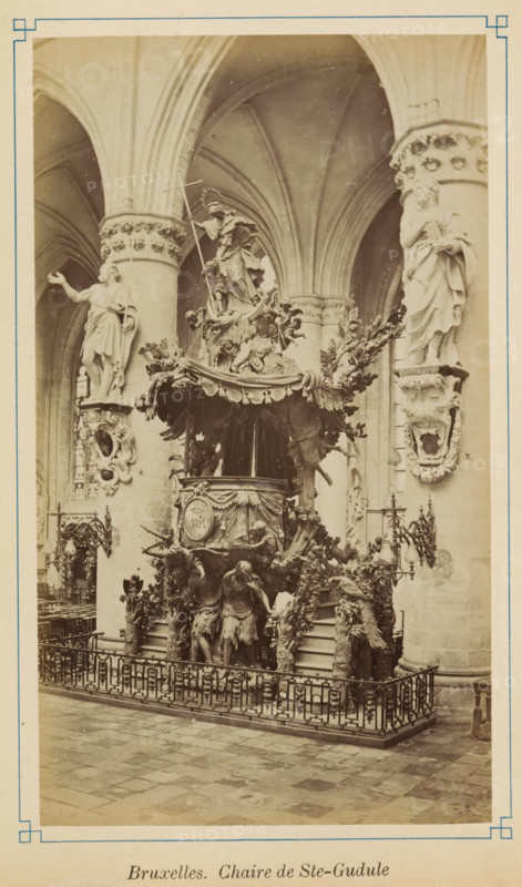 Pulpit of the Cathedral of Saint Michael and Saint Gudula in Brussels, Bruxelles. Chaire de Ste-Gudule, photograph, page, Cathedral of Saint Michael and Saint Gudula, c. 1875 - c. 1880, photographer, Brussels, paper, albumen print, height 172 mm x width 100 mm, pulpit, ambo, chancel, A tall ornate pulpit set against a vaulted interior with ribbed arches and decorated columns occupies the center of the image. The pulpit is richly carved with draped fabric motifs, foliage, and sculpted figures, including a central statue holding a staff positioned above the canopy and several supporting figures beneath it. Two large sculpted figures are mounted on the nearby columns flanking the pulpit, each standing on decorative corbels. Wrought iron railing surrounds the pulpit area, and patterned stone flooring fills the foreground. Wall-mounted lamps and additional carved reliefs and ornamental details are visible on the surrounding columns and walls.
