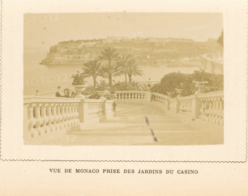 View of Monaco, seen from the gardens of the Monte Carlo casino, Vue de Monaco Prize des Jardins du Casino, photograph, page, Monaco, c. 1873 - in or before c. 1878, photographer, paper, albumen print, albumendruk, height 59 mm x width 89 mm, garden, prospect of city, town panorama, silhouette of city, A faded sepia photograph shows a broad stone staircase with ornate balustrades on both sides leading down toward a terrace lined with large urn planters and leafy shrubs; several tall palm trees rise beyond the terrace in front of a calm body of water, and across the water a rocky promontory with clustered buildings sits beneath a hazy sky.