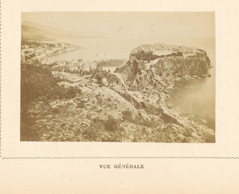 View of Monte Carlo in Monaco, Vue Générale, photograph, page, Monte Carlo, c. 1873 - in or before c. 1878, photographer, paper, albumen print, height 59 mm x width 88 mm, prospect of city, town panorama, silhouette of city, A sepia-toned aerial view shows a coastal landscape with rugged cliffs descending to a calm sea, a fortified structure perched atop a promontory with visible walls and towers, clusters of buildings and roads nestled among trees on the slopes, a bay curving into the distance with low hills along the shoreline, and areas of vegetation and rocky terrain in the foreground.