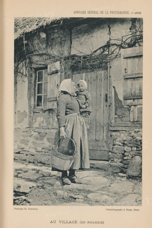 Woman with a child in her arm in a village in Picardy, France, Au Village (En Picardie), page, photomechanical print, inscription: 'ANNUAIRE GÉNÉRAL DE LA PHOTOGRAPHIE, 5e ANNÉE', photographer: Charles Commessy, Picardy, cliché maker: Jules Royer, Nancy, c. 1891 - in or before 1896, paper, collotype, photoprint, height 167 mm x width 114 mm, adult woman, tot, toddler, the ages of man, A monochrome photograph shows an older woman standing outdoors in front of a rustic wooden door and shuttered window of a weathered stone and plaster building, wearing a headscarf, a fitted long-sleeved top and a long skirt, holding a wooden bucket by its handle, and carrying a small child on her hip who is also wearing a head covering and a simple outfit; the doorway and window are framed by rough wooden beams and tangled bare vines, the ground is uneven with stones and a broom leans against the wall near a low stone pile.
