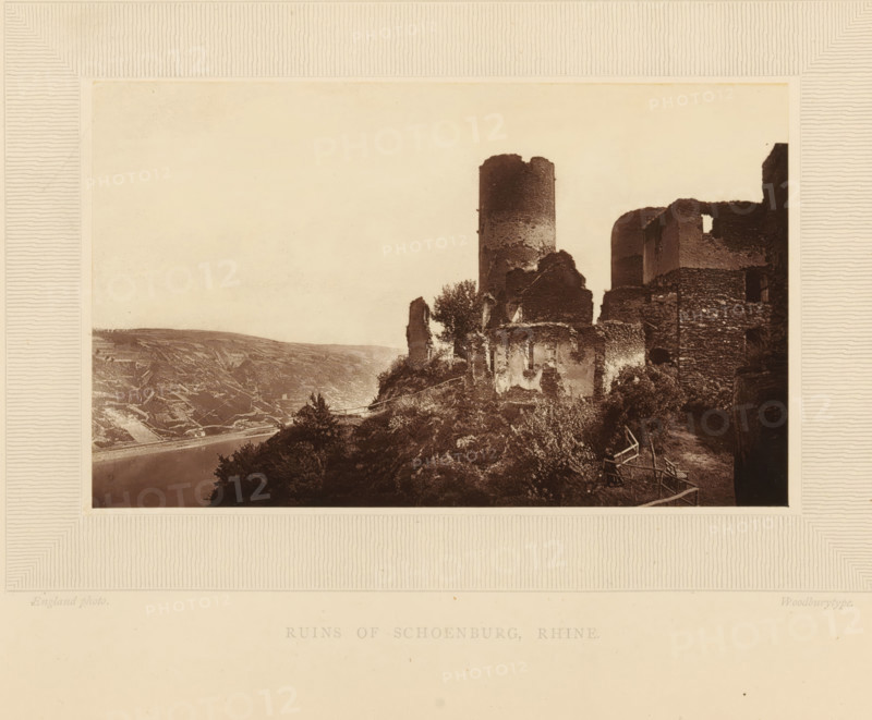Ruins of Schoenburg, Rhine, View of the ruins of Schönburg at Oberwesel, page, photomechanical print, inscription: 'England photo.', inscription: 'Europe', photographer: William England, Oberwesel, cliché maker: Woodbury Permanent Photographic Printing Company, c. 1870 - in or before 1875, Rhineland-Palatinate, paper, pasteboard, Woodburytype, Woodburytypepie, height 97 mm x width 158 mm, ruin of a dwelling, house, castle, Schönburg, A sepia-toned photograph framed by a textured mat shows a hillside with stone ruins and a cylindrical tower rising above crumbling walls, adjacent rounded battlements and fragmented masonry, vegetation growing among the stones and along the slope, a narrow path with simple wooden railings winding beside the ruins, a river visible at the base of the hill and terraced fields on the opposite bank, and a pale sky occupying the upper portion of the image.