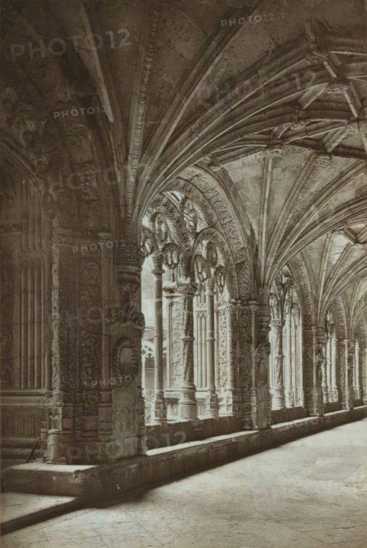 The cloisters of Belem, Cloister of the Hieronymite Monastery in Lisbon, page, photomechanical print, inscription: 'Stuart photo.', inscription: 'Europe', photographer: unknown, Mosteiro dos Jerónimos, cliché maker: Woodbury Permanent Photographic Printing Company, c. 1870 - in or before 1875, Lisbon, paper, pasteboard, Woodburytype, Woodburytypie, height 158 mm x width 106 mm, cloisters, monastery, A sepia-toned photograph shows a long covered walkway with a vaulted ceiling supported by a series of ornate stone arches and carved columns. The ceiling features ribbed vaulting with decorative bosses at the intersections, while the arches and column surfaces display intricate relief carvings of foliage and geometric patterns. Light falls through tall arched openings along one side, casting soft shadows on the smooth stone floor and the raised stone ledge that runs beside the columns. The photograph is mounted on a textured card with a patterned border surrounding the image.