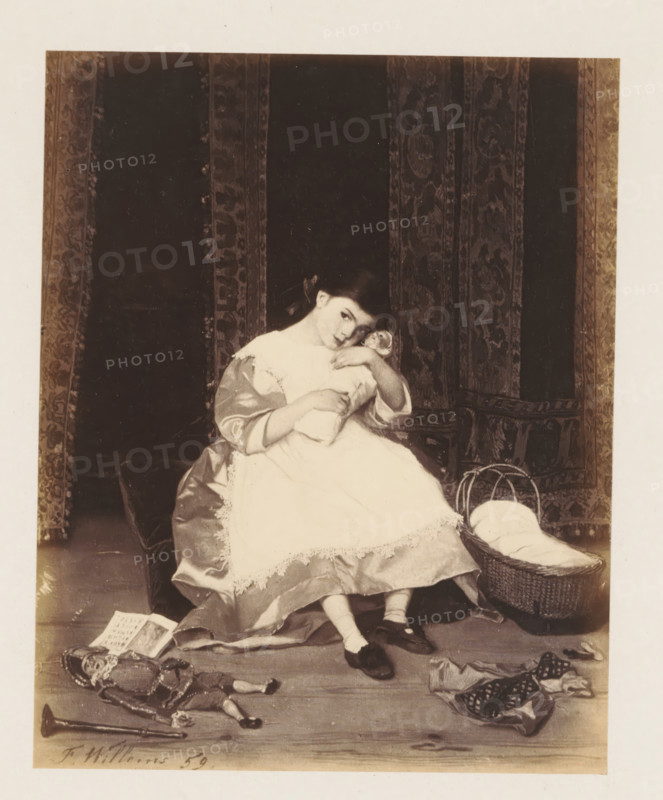 Photo reproduction of La poupée by Florent Willems, photograph, reproduction, page, after painting by Florent Willems, c. 1858 - c. 1863, photographer, paper, albumen print, albumendruk, height 141 mm x width 114 mm, picture, painting, girl (child between toddler and youth), (playing with) dolls, A seated young girl in a light-colored dress with lace trim holds a bundled doll to her chest and leans her head to one side; she wears dark shoes and light stockings and has a ribbon in her hair. She sits on a cushioned surface in front of a dark patterned curtain with vertical decorative panels. A woven basket with a cloth inside rests beside her on the floor. Scattered on the floor in front of her are a toy soldier or puppet with ornate clothing, a small musical instrument, a folded piece of fabric with patterns, and an open book. The image has a pale border around the central scene.