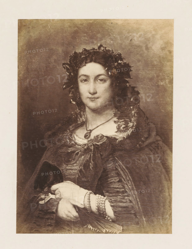 Photo reproduction of Le Domino by Jan Frans Portaels, photograph, reproduction, page, after painting by Jean François Portaels, c. 1858 - c. 1863, photographer, paper, albumen print, albumendruk, height 152 mm x width 111 mm, picture, painting, adult woman, A sepia-toned portrait of a woman shown from head to mid-torso wearing a dark, textured cloak and a leafy wreath in her dark hair, with a round pendant on a thin necklace at her throat; her face is calm with eyes looking forward and her hands rest in front of her, one gloved and the other adorned with bracelets and holding a small object, all set against a plain, slightly mottled background.