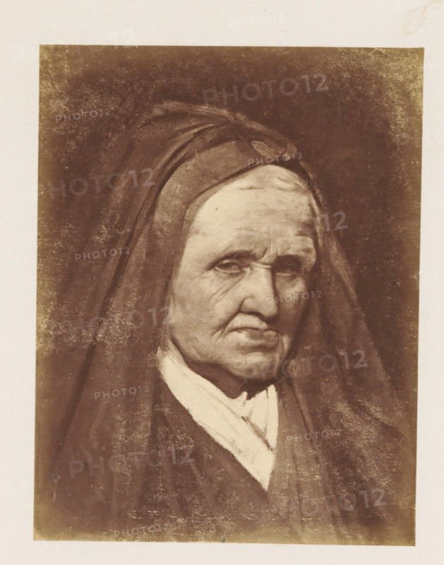 Photo reproduction of a portrait of Marie de Médicis by Paul Delaroche, photograph, reproduction, page, after painting by Paul Delaroche, c. 1858 - c. 1863, photographer, paper, albumen print, height 142 mm x width 110 mm, Person, Maria de' Medici, picture, painting, old woman, A sepia-toned portrait of an elderly person wearing a dark hooded garment and a light-colored scarf, showing a face with deep wrinkles, downturned mouth, and heavy eyelids. The hood frames the head and casts shadows across the forehead and cheeks. The background is dark and textured, and the image is bordered by a lighter margin. The clothing appears layered with visible folds and a contrast between the dark outer garment and the lighter fabric at the neck.