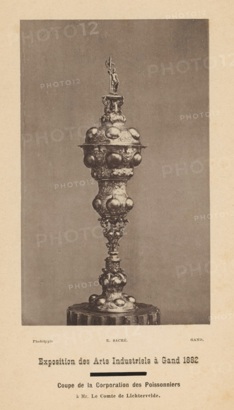 Cup de la corporation des poissonniers, Coupe de la corporation des poissonniers, page, photomechanical print, inscription: 'Exposition des Arts Industriels à Gand 1882, à Mr. Le Comte de Lichtervelde', cliché maker: Edmond Sacré, c. 1878 - in or before 1883, photographer, paper, collotype, photoprint, height 137 mm x width 85 mm, Print opposite title page, mug, beaker, goblet, A tall ornate metal chalice-like object centered against a plain background, featuring a narrow decorative stem rising from a round pedestal base with bulbous rounded protrusions and intricate embossed patterns, above which a wider section with repeated rounded bulges and elaborate relief work forms a large cup topped by a domed lid adorned with more rounded protrusions and a small sculpted figure standing on a decorative finial.