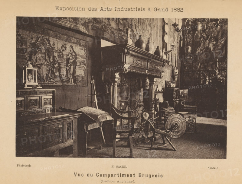 Interior of the Brussels part of the Exposition des Arts Industriels, Vue du compartment Brugeois (section ancienne), page, photomechanical print, inscription: 'Exposition des Arts Industriels à Gand 1882', cliché maker: Edmond Sacré, c. 1878 - in or before 1883, photographer, paper, collotype, photoprint, height 101 mm x width 152 mm, book - MM - book open, open hearth, fire-place, stool, A sepia-toned photograph of a room filled with ornate antique furniture and decorative objects, including a large carved wooden mantel with vases and pitchers arranged on top, a tapestry or painted wall hanging depicting robed figures, a wooden cabinet with carved panels, a cushioned bench, and a spinning wheel among various chairs and small tables; chandeliers and wall-mounted plates are visible on the right, and smaller decorative items and framed pieces sit on surfaces throughout the space.