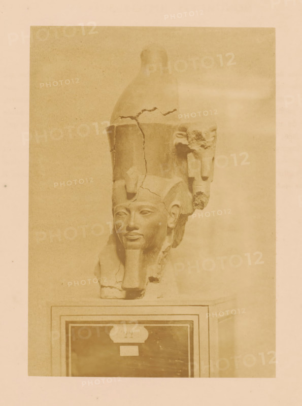 Head of an Egyptian statue in the Boulaq Museum in Cairo, Egypt, photograph, page, photographer: Hippolyte Délié, Cairo (city), photographer: Emile Béchard, c. 1866 - in or before 1871, Ottoman Empire, North Africa, paper, albumen print, height 202 mm x width 142 mm, permanent exhibition, museum, piece of sculpture, reproduction of a piece of sculpture (Egyptian art), Boulaq Museum, A sepia-toned photograph of a carved stone bust mounted on a rectangular plinth, showing a human face with a stylized beard and a tall headdress that is cracked and partially broken, with fragments and surface damage visible on the headdress and side area, the bust centered within a lighter rectangular frame against a plain background, the plinth featuring a framed panel with contrasting darker interior and lighter border.